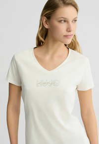 T-shirt blanc à manches courtes avec un col en V et un logo décoratif en strass sur la poitrine. Matière en coton doux avec une coupe décontractée.