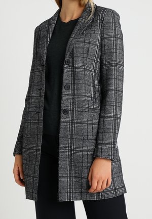 Frau, die einen knielangen grauen karierten Blazer mit drei Knöpfen über einem schwarzen Oberteil und einer schwarzen Hose trägt, steht vor einem schlichten Hintergrund.