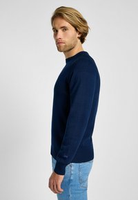 Granatowy sweter dziany z okrągłym dekoltem, ściągaczami na mankietach i dolnym hemie, z dyskretnym logo na nadgarstku. Tkanina wydaje się gładka i miękka.