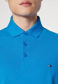 Blauwe polo gemaakt van structuurstof, met een klassieke kraag, drie knopen en een klein rood, wit en blauw logo op de borst.