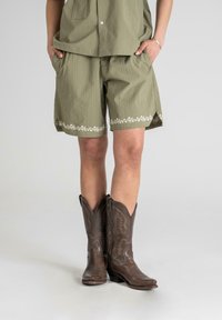 Olivenfargede shorts med stripete tekstur, med blomsterbroderi langs kanten, sammen med brune lær cowboy støvler med intrikate mønstre.