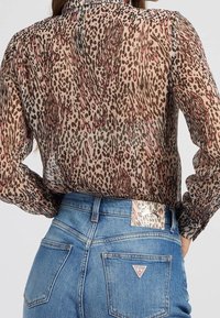 Bluse med leopardprint stoppet ned i højtsiddende blå jeans med synligt Guess-logo på baglommen og mærket på taljebåndet.
