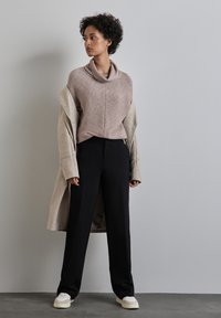 Maglione beige a maglia con collo a cascata, indossato sotto un lungo cappotto beige. I pantaloni neri a gamba dritta e le sneaker color crema completano l'outfit.