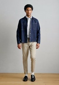 Σκούρο μπουφάν τζιν με πλαϊνές τσέπες, λευκό πουκάμισο με γιακά, μπεζ στενά παντελόνια και μαύρα μοκασίνια, πάνω σε ανοιχτό γκρι φόντο.