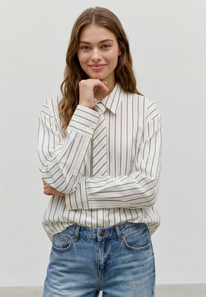 Jeune femme aux longs cheveux ondulés portant une chemise blanche à rayures et un jean bleu, souriant avec une main posée sur le menton.