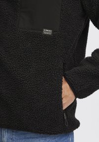 Svart fleece-pullover med en teksturert, plysjaktig overflate, som har en ensfarget svart brystlomme og en liten merkeetikett. Inkludert sidelomme med glidelås.