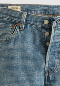 Jeans de denim azul claro con un acabado desgastado, que cuentan con cinco botones y costuras visibles. La tela presenta una textura clásica de canalé.