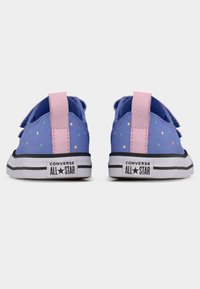 Ζευγάρι μοβ αθλητικών παπουτσιών Converse All Star με πολύχρωμες πουά βούλες και ροζ ταμπελάκια στη φτέρνα, εμφανίζεται από πίσω σε λευκό φόντο.