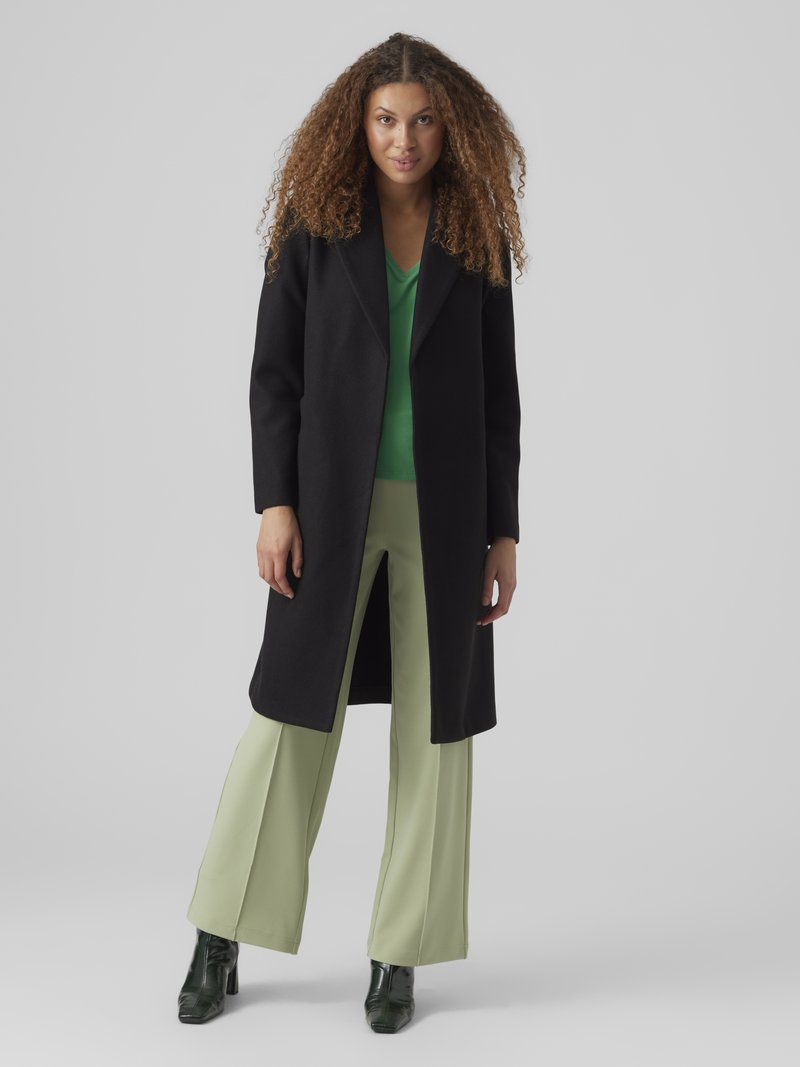 Sort lang frakk med hakket krage, kombinert med en grønn skjorte og vide, lysegrønne bukser. Glinsende svarte ankelstøvler fullfører antrekket.