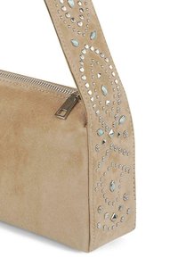 Bolso de mano de ante beige con una correa decorativa de costura, adornos metálicos y similares a gemas, cierre con cremallera y textura suave.