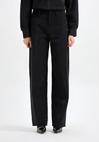 Zwarte wijde broek met een gestructureerd oppervlak, voorzien van subtiele schitteringen, voorzakken en een hoge taille. Glanzende schoenen maken de look compleet.