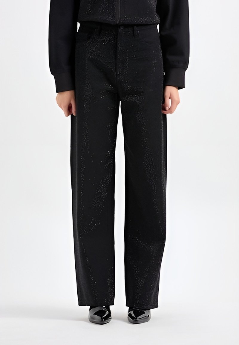 Zwarte wijde broek met een gestructureerd oppervlak, voorzien van subtiele schitteringen, voorzakken en een hoge taille. Glanzende schoenen maken de look compleet.