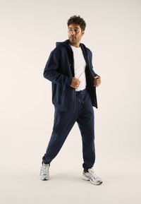 Mørkeblå treningstopp med hette og joggebukser laget av myk stoff, med glidelås foran, snor i livet, og sneakers med sølvdetaljer.