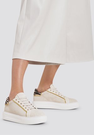 Beige Sneaker mit strukturierter Oberfläche, goldenen Akzenten und schwarz-weiß gestreiften Details an der Ferse, auf einer weißen Sohle.