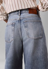 Άτομο που φοράει φωτεινό μπλε τζιν ψηλόμεσο και πουκάμισο με μακριά μανίκια με καφέ κάθετες ρίγες, μπαλωμένο μέσα, σε ουδέτερο φόντο.