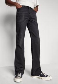 Jeans de mezclilla negros acampanados con una textura ligeramente desgastada, diseño de cinco bolsillos y cierre de botón, llevados con zapatillas negras de corte bajo.