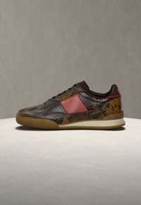 Παπούτσι τύπου sneaker χαμηλού προφίλ με καφέ υφή φιδιού, σκούρα πράσινα κορδόνια, κόκκινο πλαϊνό πάνελ και ανοιχτό καφέ σόλα σε ουδέτερο φόντο.