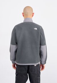 Persona con la testa rasata che indossa una giacca in pile grigia con il logo "The North Face" sulla parte superiore della schiena, abbinata a pantaloni cargo neri.