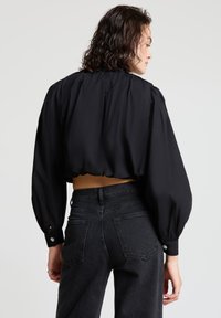 Schwarze, kurz geschnittene Bluse mit Puffärmeln und hohem Ausschnitt, kombiniert mit schwarzen, hoch taillierten Jeans mit Gesäßtaschen und Knopfakzenten.