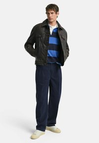 Bruine leren jasje met knoopsluiting, blauw en marinegestreept poloshirt, marineblauwe pantalons en lichtgekleurde sneakers.