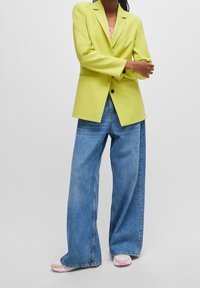 Neonowa zielona marynarka o dopasowanym kroju, z pojedynczym zapięciem na guzik, zestawiona z luźnymi, niebieskimi dżinsami oraz jasnymi trampkami.