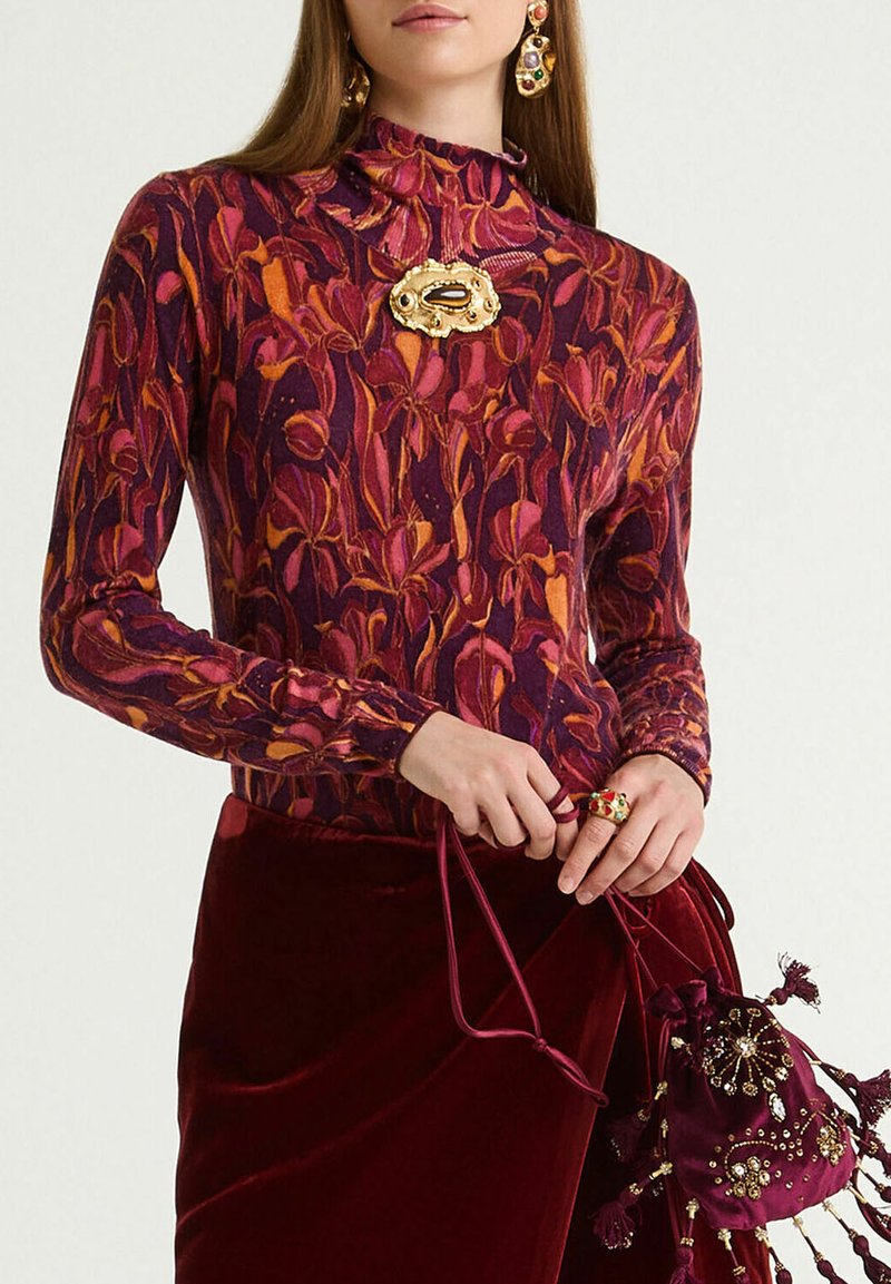 Top a maniche lunghe con motivo floreale viola e arancione, caratterizzato da un collo alto e una spilla decorativa dorata. Abbinalo a una gonna di velluto color bordeaux.