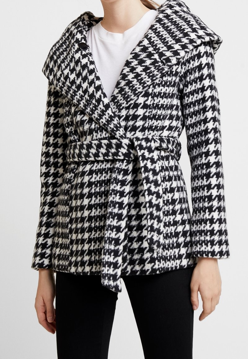 Cappotto houndstooth bianco e nero con ampio colletto, cintura a cravatta e maniche lunghe, realizzato in un tessuto testurizzato.