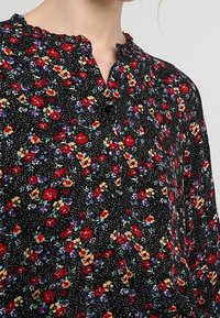 Svart blommig blus med röda och gula blommor. Har rundad halsringning, knapplist och textil med strukturerat tyg med svarta prickar.