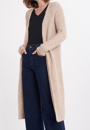 Žena v dlouhém béžovém pleteném kardiganu přes černý top s výstřihem do V a tmavě modrých širokých džínech s vysokým pasem, s rukama v kapsách.