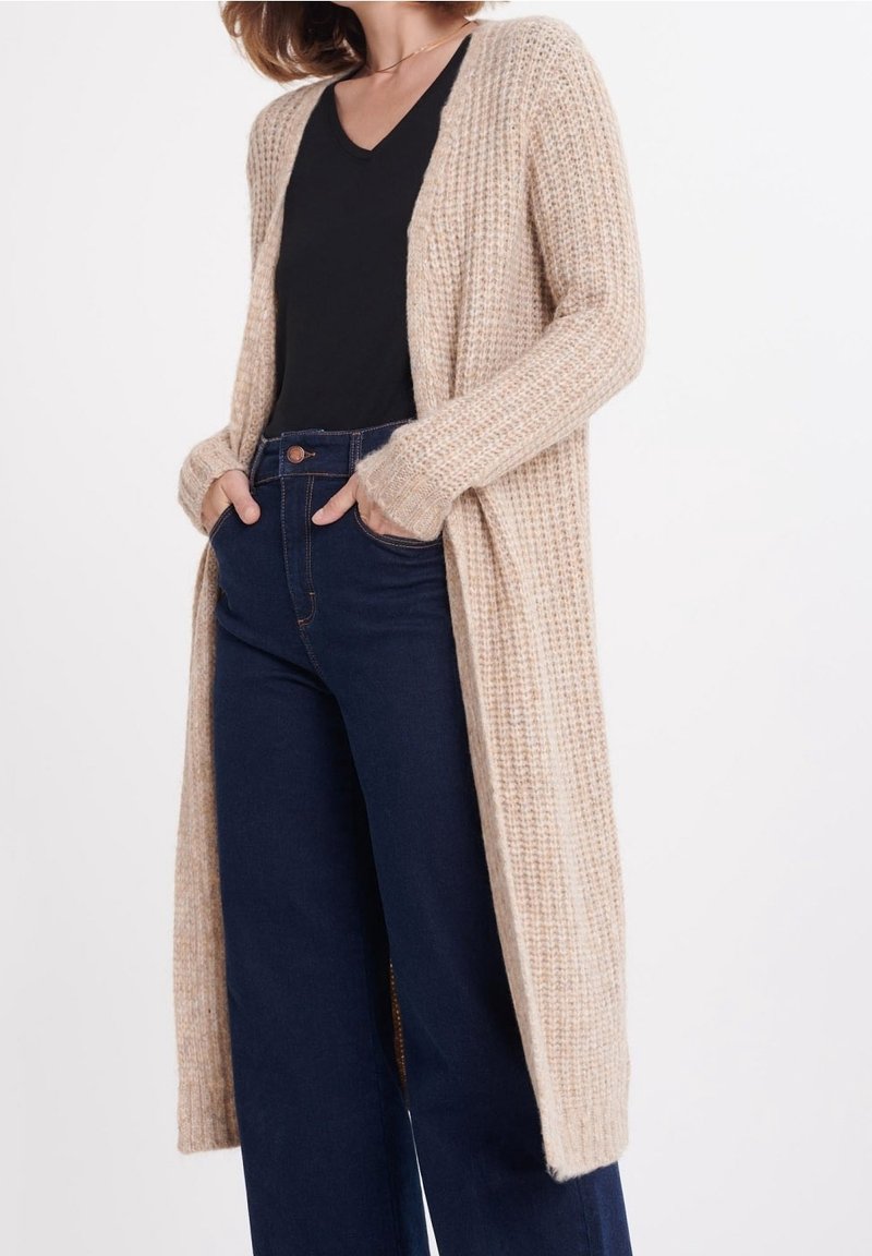 Žena v dlouhém béžovém pleteném kardiganu přes černý top s výstřihem do V a tmavě modrých širokých džínech s vysokým pasem, s rukama v kapsách.