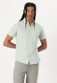 Camicia a maniche corte, di colore verde chiaro, realizzata in materiale morbido, con patta dei bottoni e una vestibilità casual. Indossata con pantaloni in denim chiari.