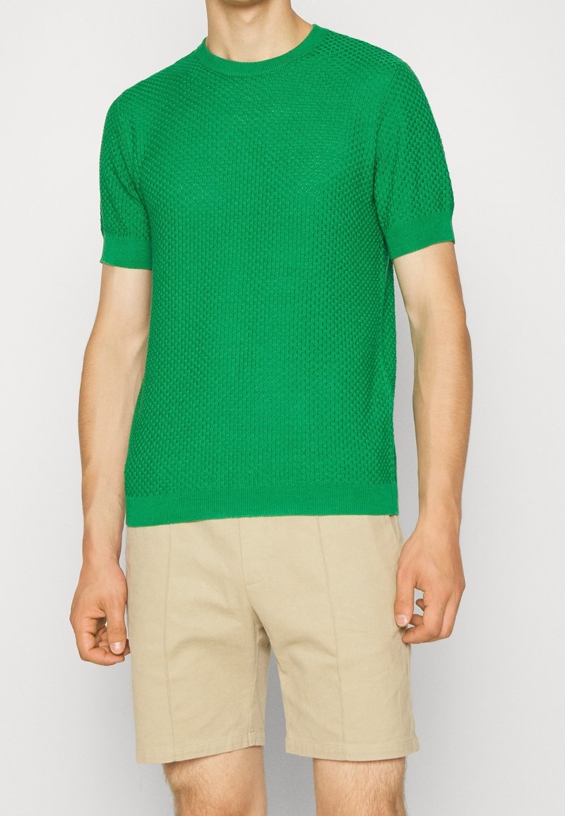 Grön kortärmad stickad tröja med strukturerad mönster och ribs i kragen, kombinerad med beige shorts.