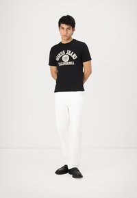 Camiseta de algodón negra con el logo "Guess Jeans California" en impresión blanca y dorada, combinada con pantalones blancos y zapatos negros.