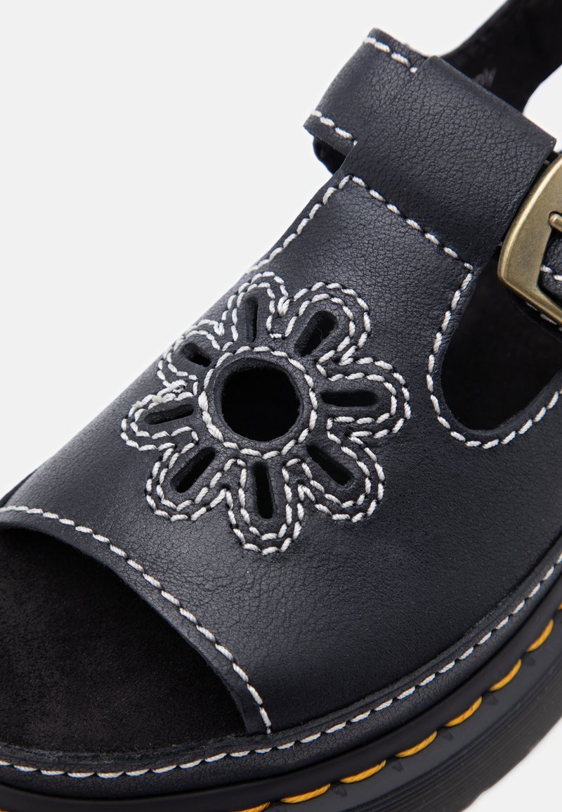 Sandalo in pelle nera con cuciture bianche, intaglio a forma di fiore sulla parte superiore, punta aperta e fibbia in ottone sulla cinghia alla caviglia.