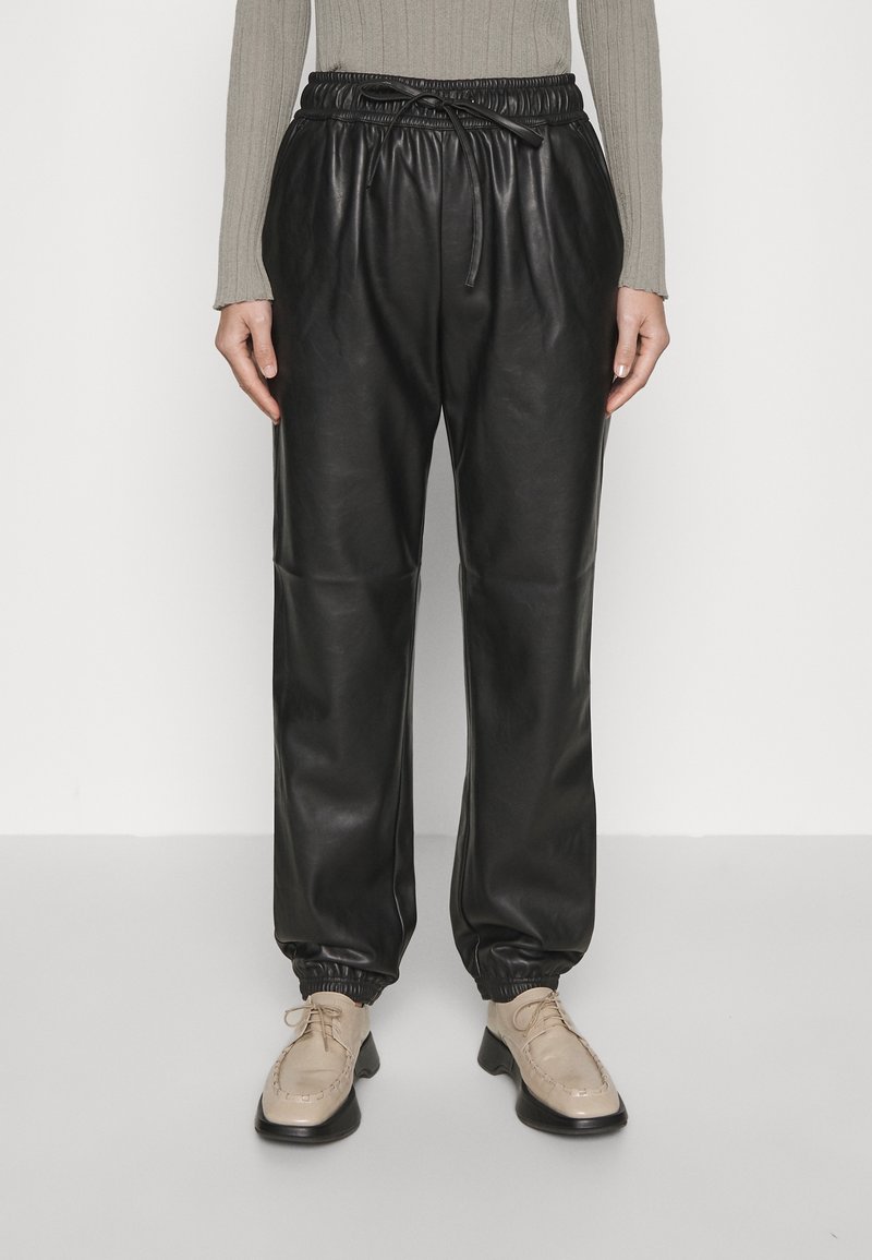 Personne portant un pantalon jogger en similicuir noir avec des poignets élastiques et des chaussures beiges à lacets, debout devant un fond blanc uni.