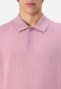 Světle růžové polo tričko vyrobené z texturované tkaniny, s límečkem, třemi knoflíky a čistým designem bez vzorů nebo akcentů.