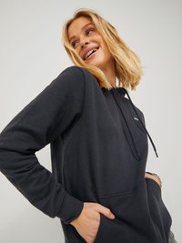 Felpa nera con cappuccio a forma di kangaroo, realizzata in tessuto morbido, dotata di cappuccio con stringhe e polsini a costine. Modello in posa con un sorriso rilassato.