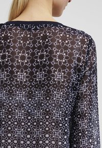 Przezroczysta czarna bluzka z delikatnym białym wzorem kwiatowym, z okrągłym dekoltem i długimi rękawami, podkreślająca misternie zaprojektowane detale.