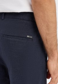 Granatowe spodnie z teksturowanym, kratkowanym wzorem. Posiadają bezpieczną tylną kieszeń oraz metalową metkę z logo marki. Prosty krój z dopasowanym pasem.