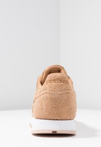 Bakvy av en beige texturerad mockaskor med vit sula och vadderad krage på en vit yta mot en ljusgrå bakgrund.