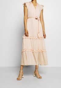 Dlouhé, světle růžové šaty s volánovými rukávy, knoflíkovým zapínáním vpředu a páskem v pase. Vícevrstvý design s volánky podél spodního okraje, splývavá látka.