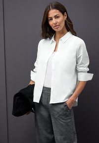 Chemise blanche à manches retroussées portée sur un haut blanc. Assortie à un pantalon à carreaux gris avec des accents métalliques. Fond sombre.