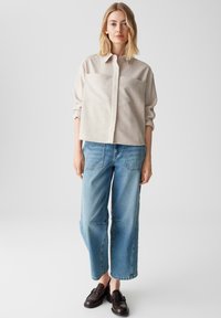 Camicia beige a maniche lunghe con bottoni, dal taglio morbido e due tasche sul petto, abbinata a jeans azzurri a gamba larga e mocassini scuri.