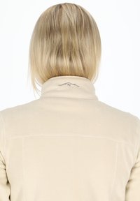 Beige fleece-takk med høy krage, med et subtilt fjelldesign brodert på baksiden av nakken. Glatt tekstur, enkelt design.