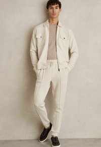 Training ușor de culoare crem, realizat dintr-un material moale, având o jachetă cu fermoar și două buzunare pe piept, precum și pantaloni cu snur și dungi laterale. Adidași negri.