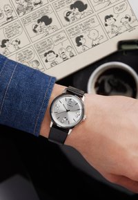 Silbernes Zifferblatt mit schwarzem Lederband. Mit Cartoonfiguren, silbernen Stundenmarkierungen und einem polierten Gehäuse. Getragen an einem Handgelenk mit Jeans.