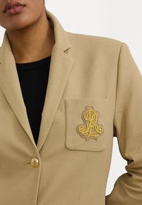 Blazer beige con tessuto testurizzato, singolo bottone dorato e tasca a toppa con un logo ricamato in oro ornamentale. Top nero sotto.