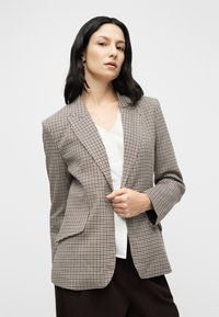 Blazer à carreaux en tissu marron, crème et noir, avec une coupe ajustée, un col à revers et des poches avant, porté sur un haut blanc.