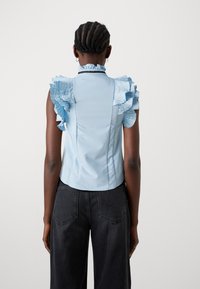 Gaiši zilas bez piedurknēm blūze ar viļņotiem plecu akcentiem, gluda tekstūra, pieguļošs dizains un augsts apkakle; valkāta ar augsta vidukļa melnajām džinsām.