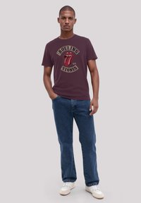 Maroon cotton t-shirt featuring a graphic of the Rolling Stones logo in red and black. Paired with blue denim jeans and white sneakers.